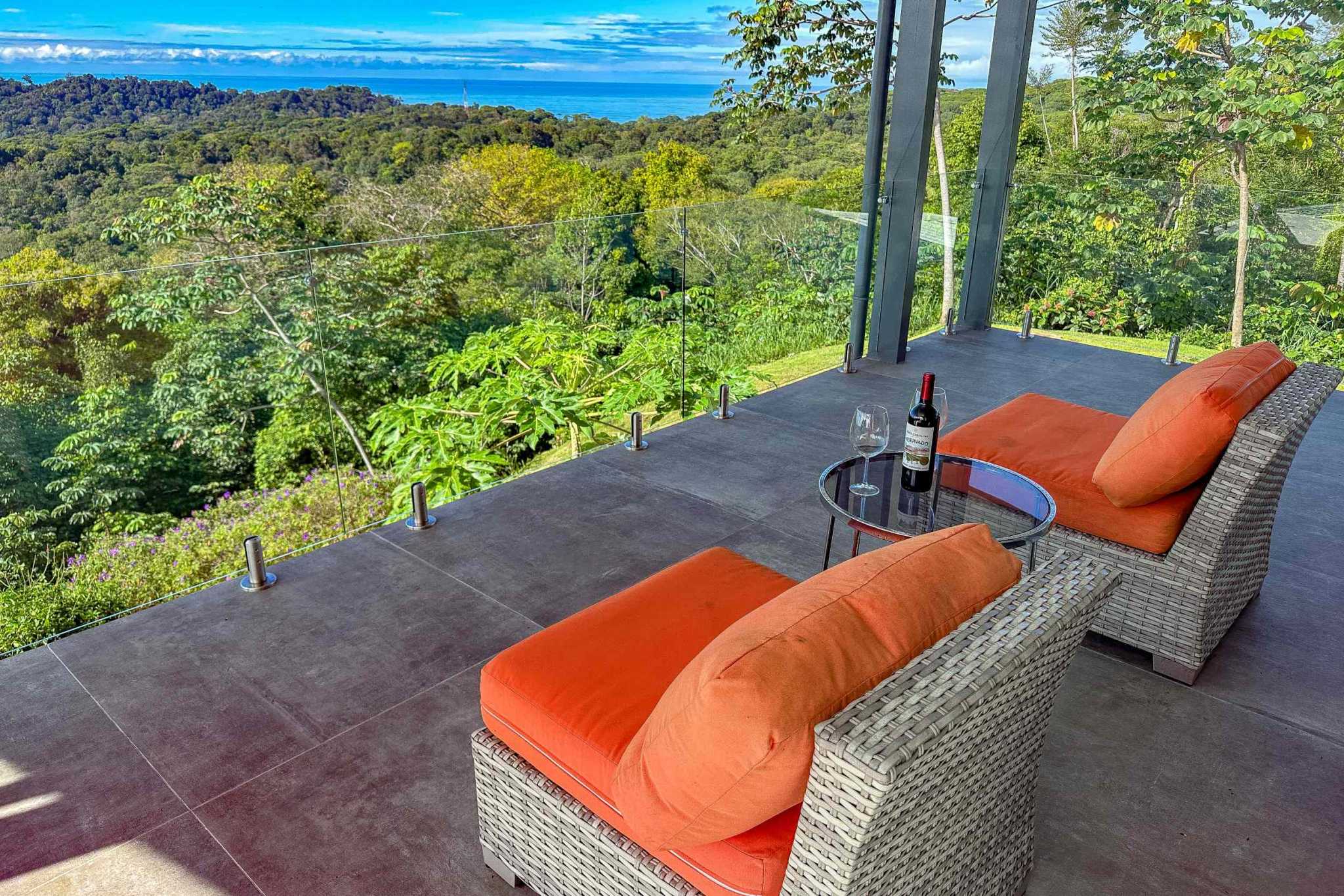 Mountain & Ocean View Retreat covered terrace with orange cushioned chairs, glass railing, jungle and ocean view in Ojochal, Costa Rica