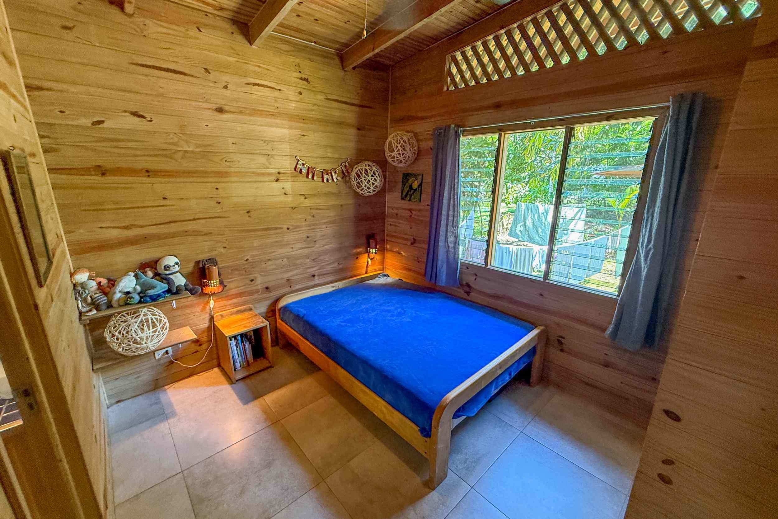 Cozy Wooden Master Bedroom Jungle Hotel wood-paneled bedroom with blue bed and forest-view window in Ojochal, Costa Rica
